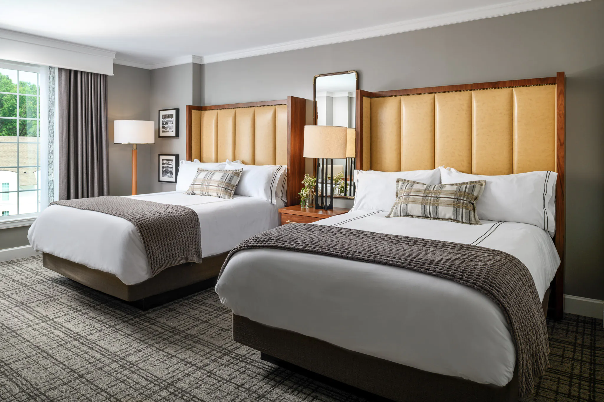 Deluxe room with two queen beds featuring amber upholstered headboards, white bedding with plaid throws, walnut nightstands, and large windows with natural light