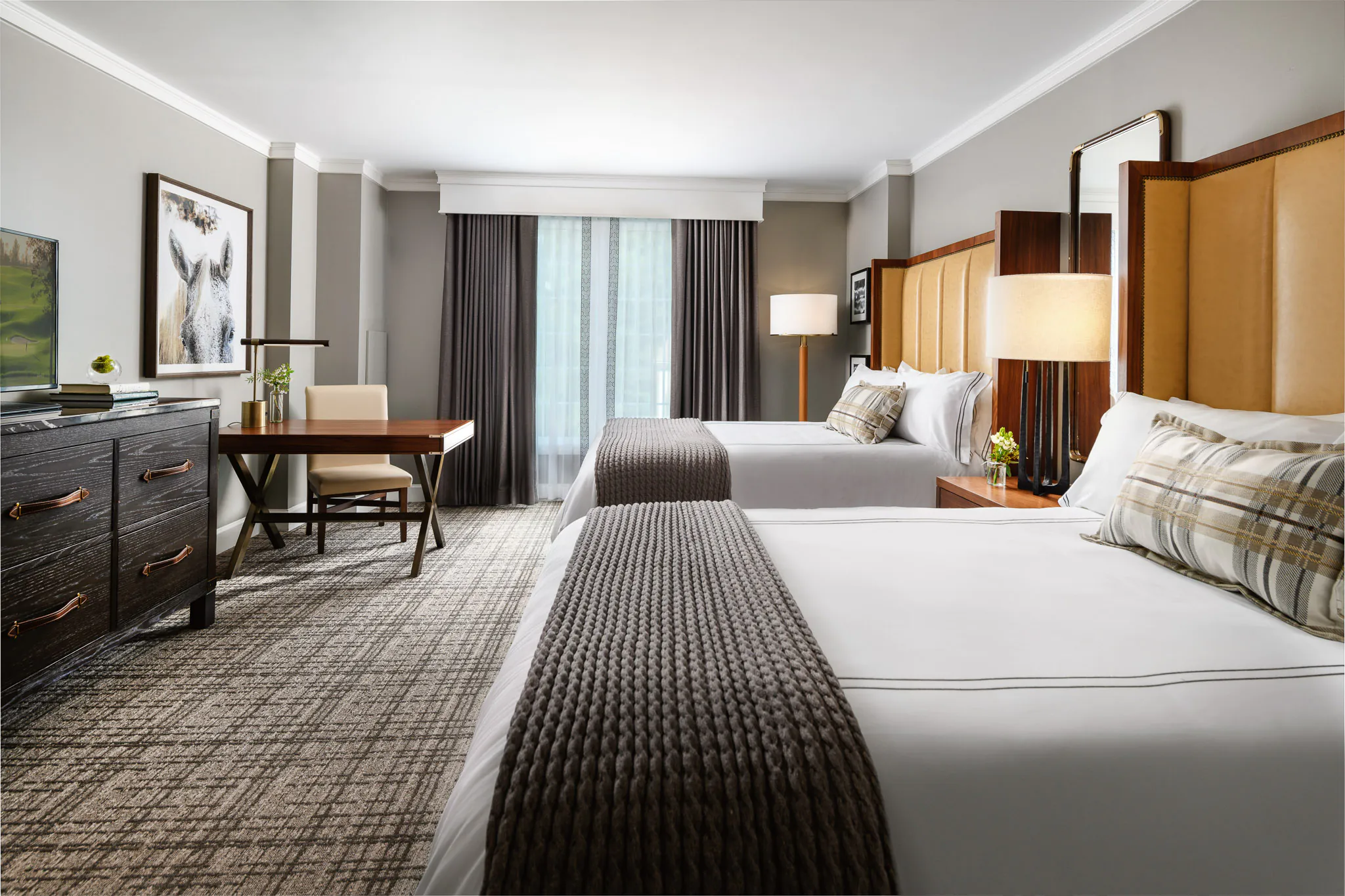Spacious Deluxe King room with amber tufted headboard, white bedding, dark wood furnishings, writing desk, and framed artwork on grey walls