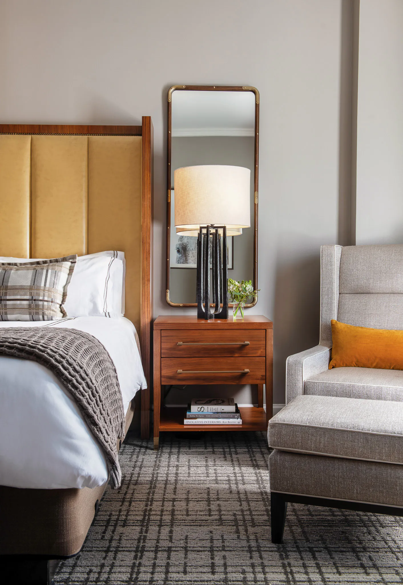 Bright Deluxe King room with amber upholstered headboard, white bedding, tufted leather bench, dark wood furnishings, and French doors with natural light