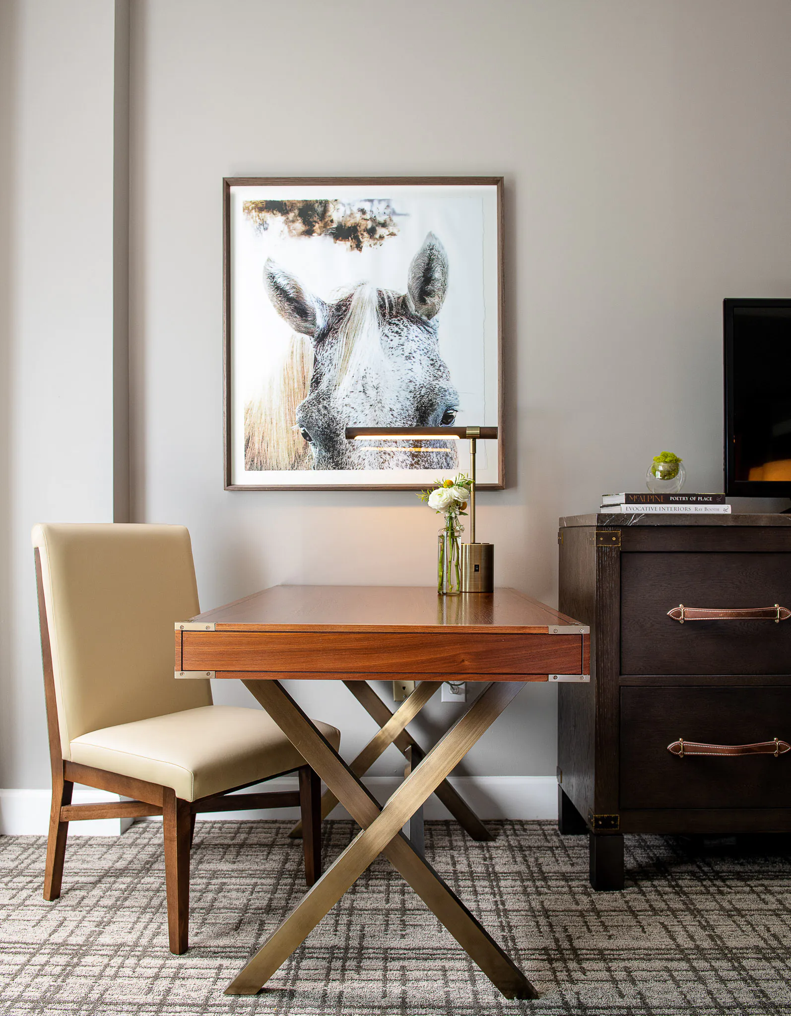 Deluxe King room workspace with X-leg writing desk, cream upholstered chair, dark wood dresser, and framed rhinoceros artwork on grey wall