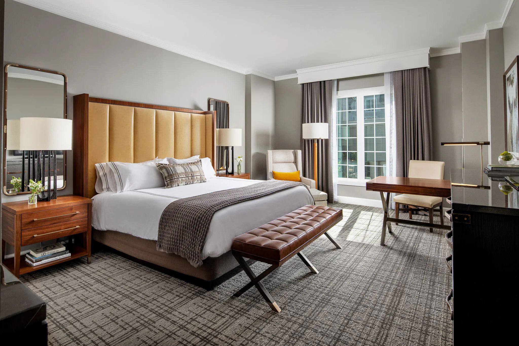 Deluxe King guest room featuring an upholstered amber headboard, white bedding with plaid throw, matching walnut nightstands with table lamps, and framed mirrors on a warm grey wall