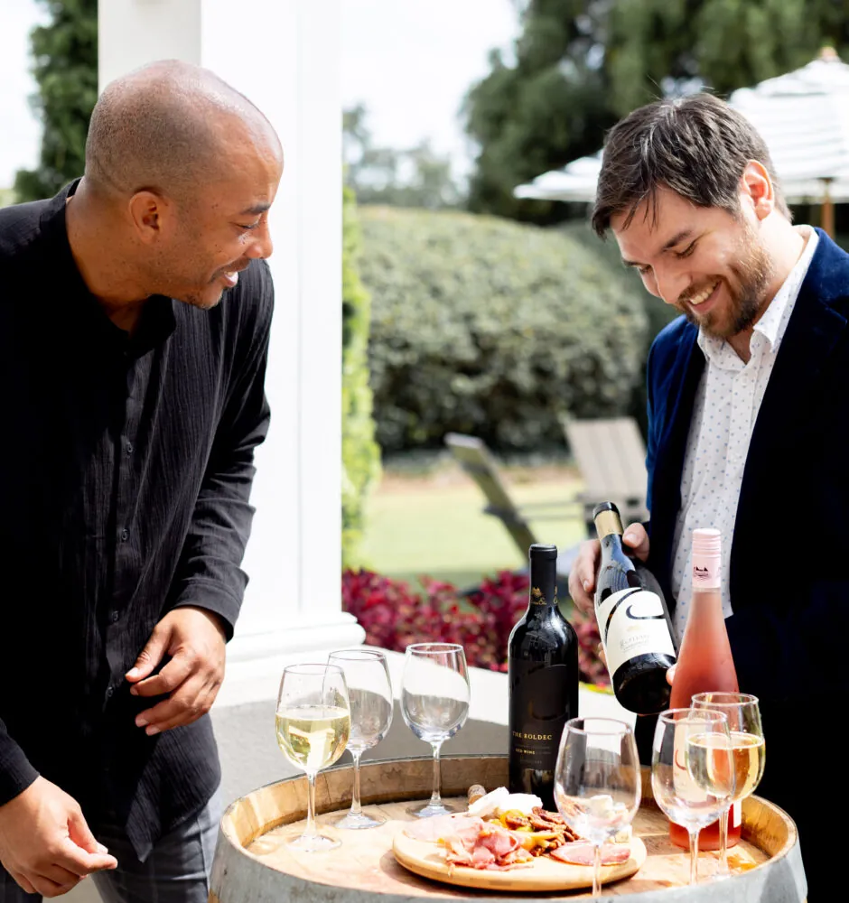 A blurry photo of two men standing on a patio, looking at a wine tasting setup on a small wooden barrel. The man on the right, wearing a blue suit jacket, is holding a bottle of rosé wine. He is smiling and looking down at the selection of wine glasses and a wooden board with snacks. The man on the left, in a black shirt, is looking at him and also smiling.