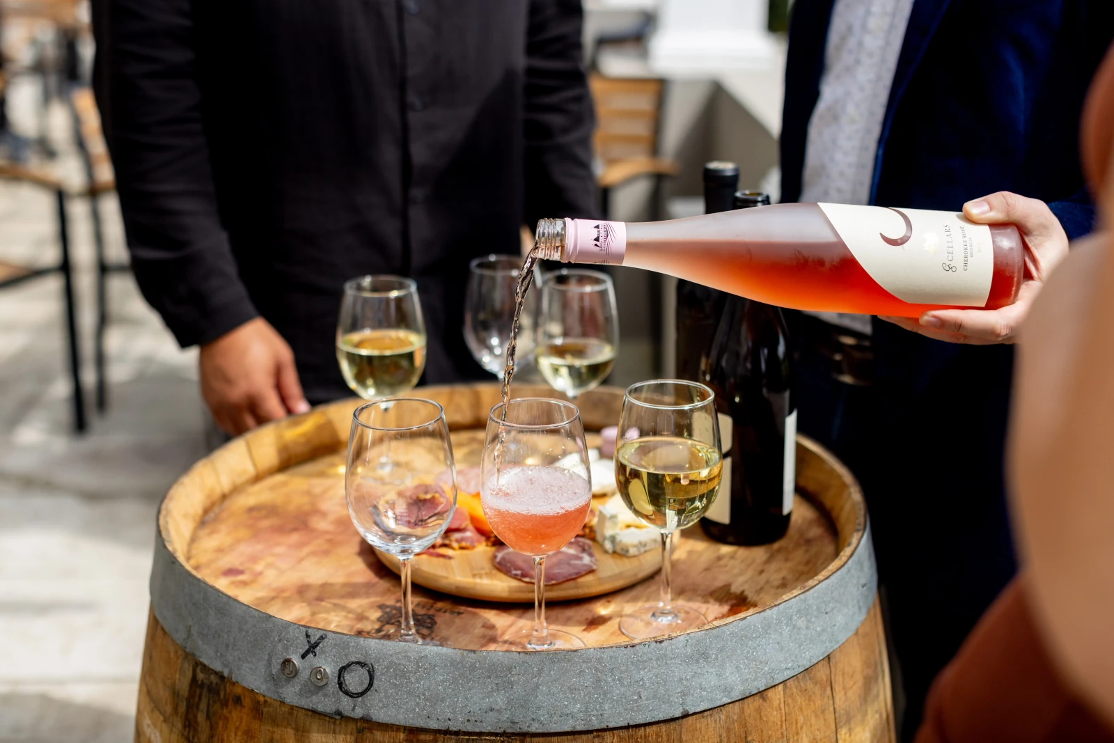 A close-up shot of a wine tasting. A person in a black jacket is pouring rosé wine from a bottle into a glass on top of a small wooden barrel. There are other glasses of white wine and a bottle already on the barrel.