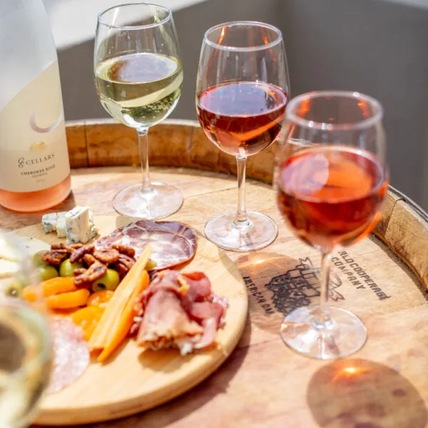 A wooden board with snacks, including meats and grapes, is on a wooden table with several glasses of wine.