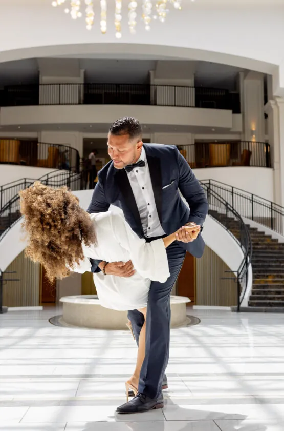 A man in a tuxedo dips a woman while dancing in a large, elegant lobby with a grand staircase.