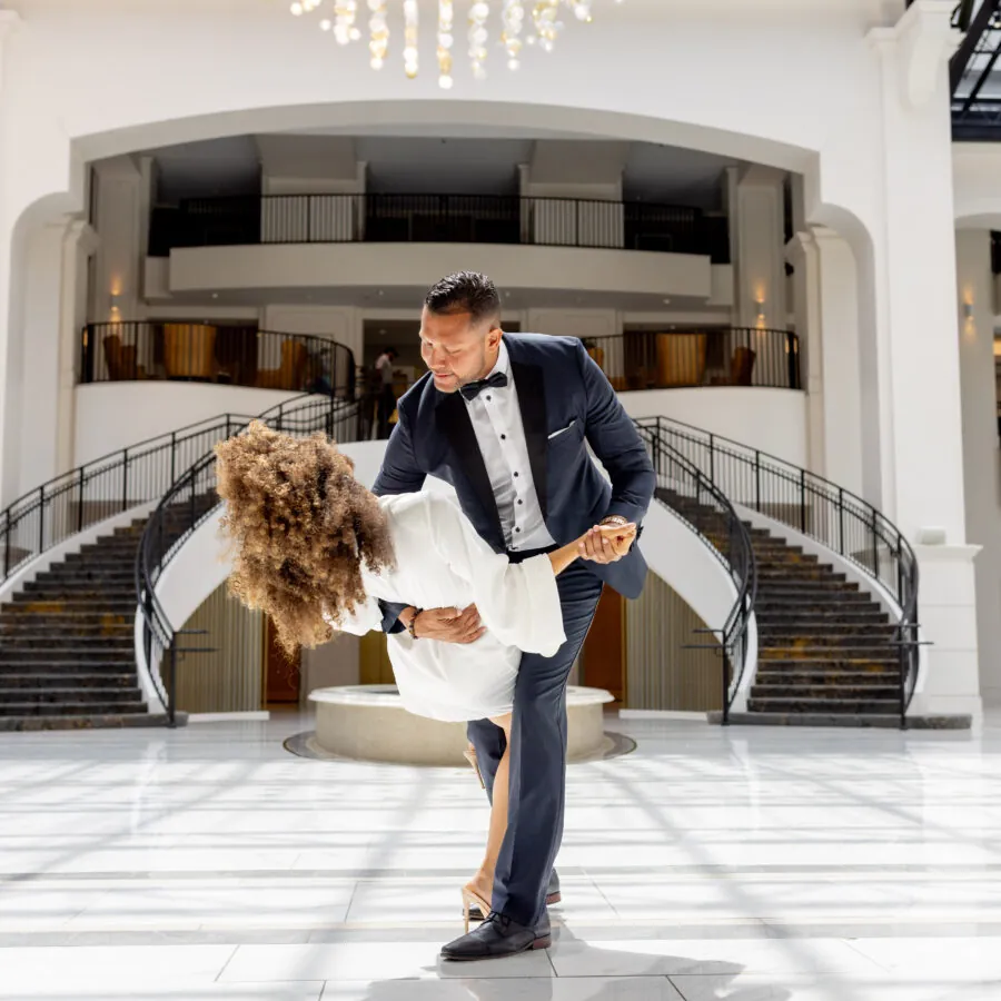 A man in a tuxedo dips a woman while dancing in a large, elegant lobby with a grand staircase.