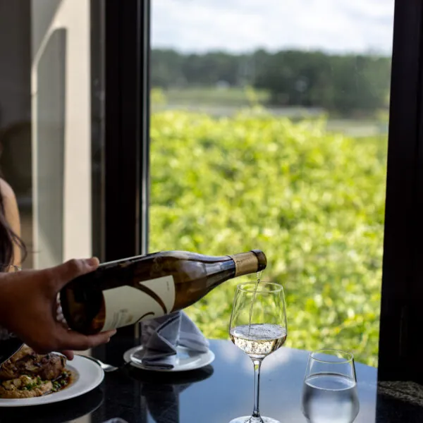 A table is seen indoors next to a window overlooking a vineyard, with plates of food and glasses on it.