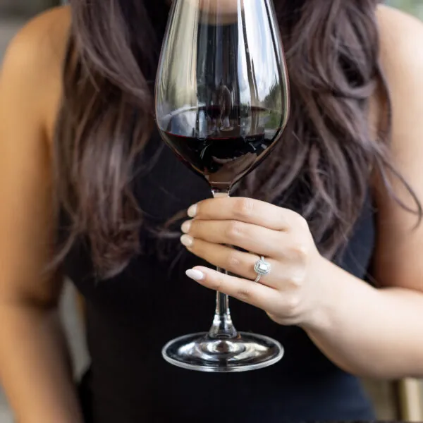 A woman is shown from the chest up, with her head bent slightly and her eyes closed as she smells a glass of red wine.