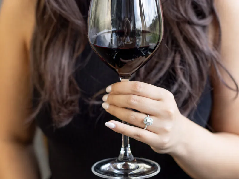 A woman is shown from the chest up, with her head bent slightly and her eyes closed as she smells a glass of red wine.