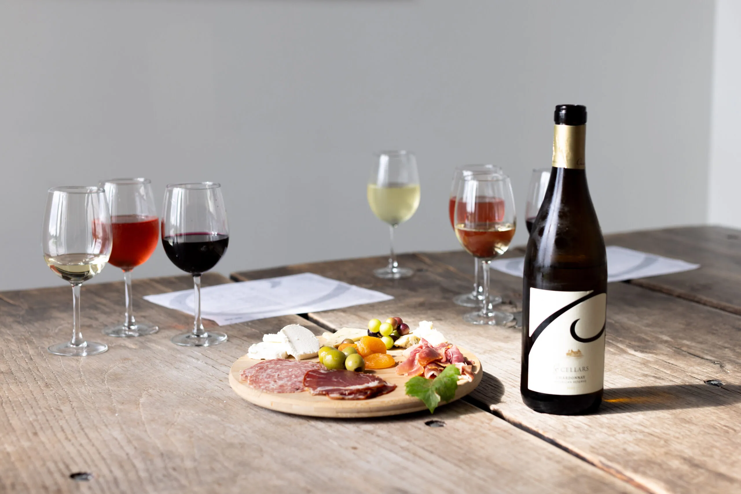 A still life of a wine tasting setup on a rustic wooden table. In the foreground, there is a round wooden board with a selection of snacks, including cheese, olives, and cured meats. A bottle of white wine stands next to the board. In the background, there are five glasses with varying amounts of white, rosé, and red wine.