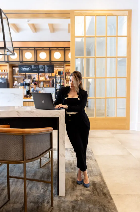 A woman in black leans on a counter, working on a laptop in a stylish, modern interior space.