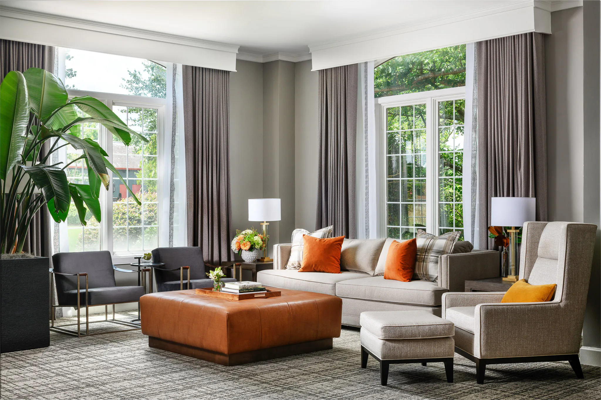 Bright suite living area with grey sofa, orange accent chairs and ottoman, floor lamp, and large windows with garden views