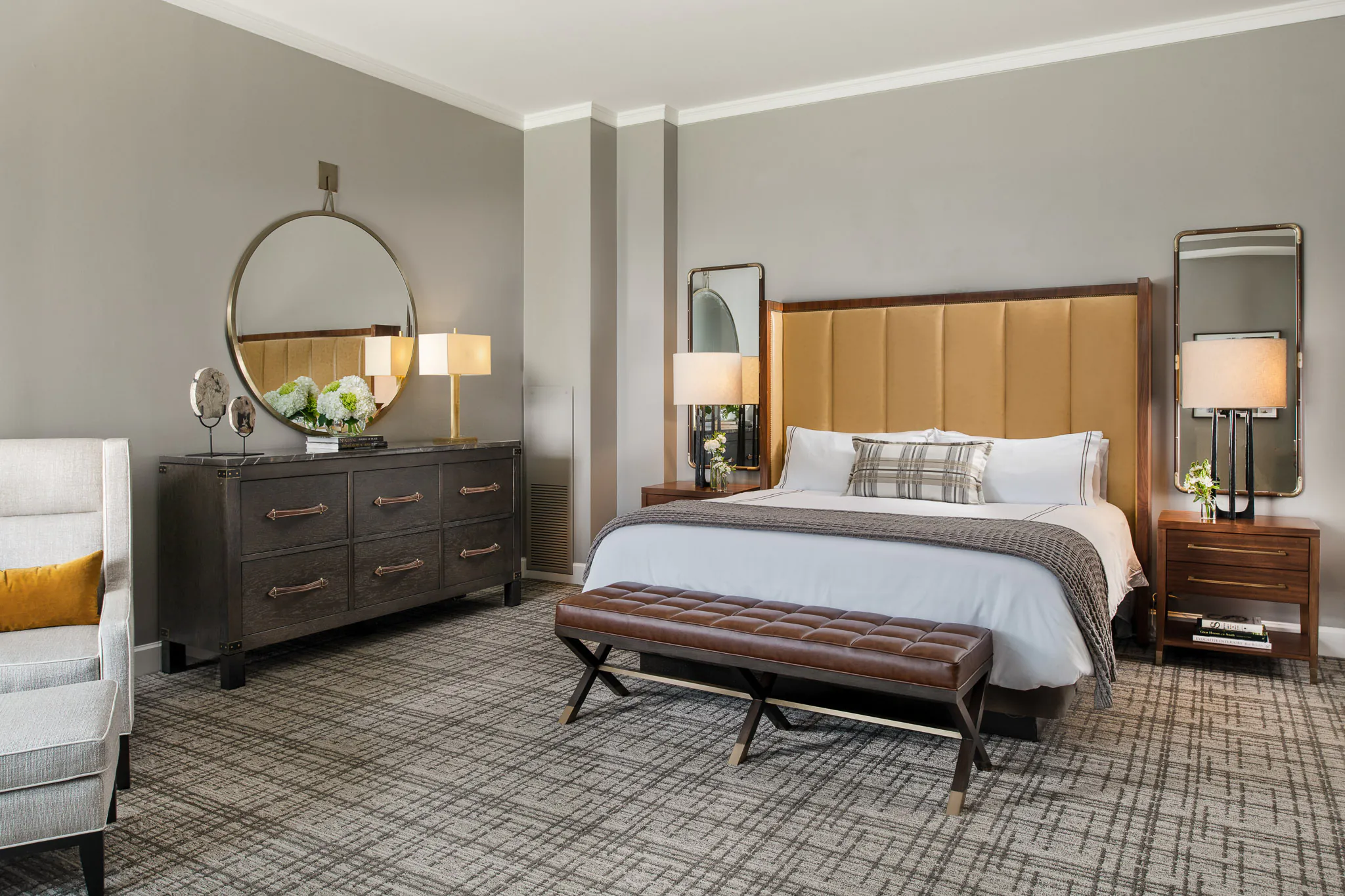 Deluxe King room with upholstered headboard, white bedding, tufted leather bench, dark wood dresser, and round mirror