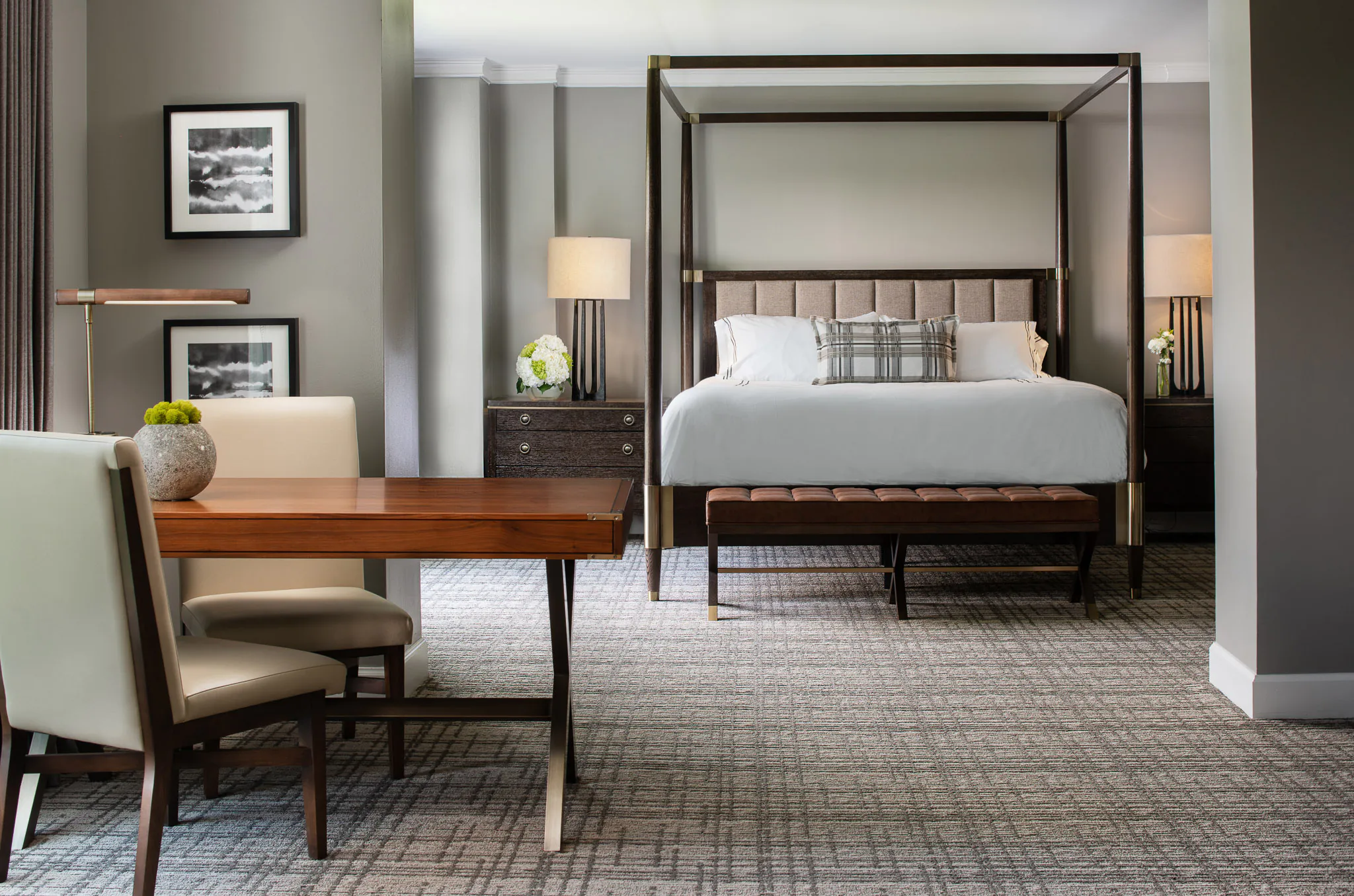 Elegant bedroom with four-poster king bed, white bedding, plaid accent pillow, wooden bench, and neutral grey walls