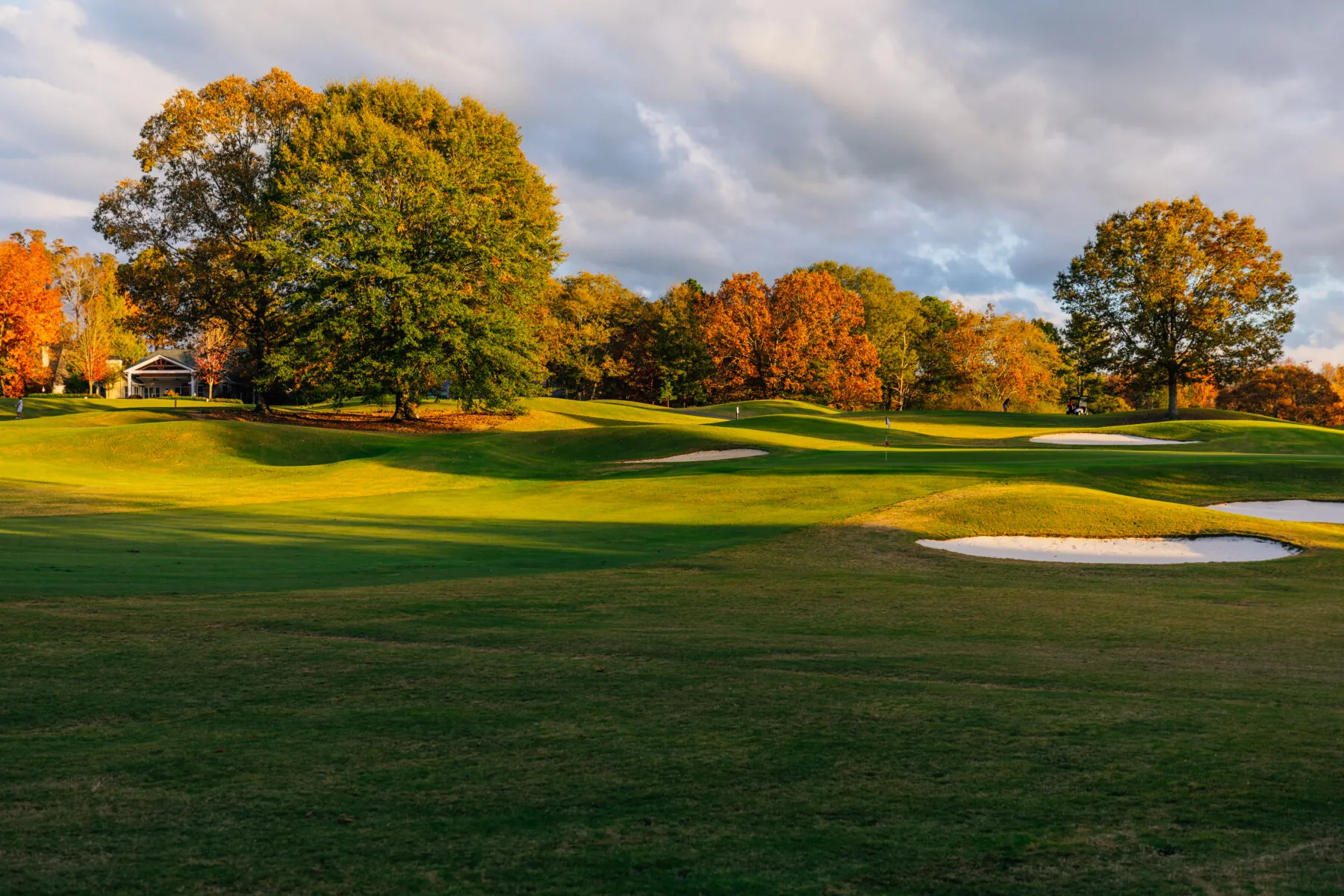 A scenic golf course with rolling green hills and trees with autumn foliage under a dramatic, cloudy sky.