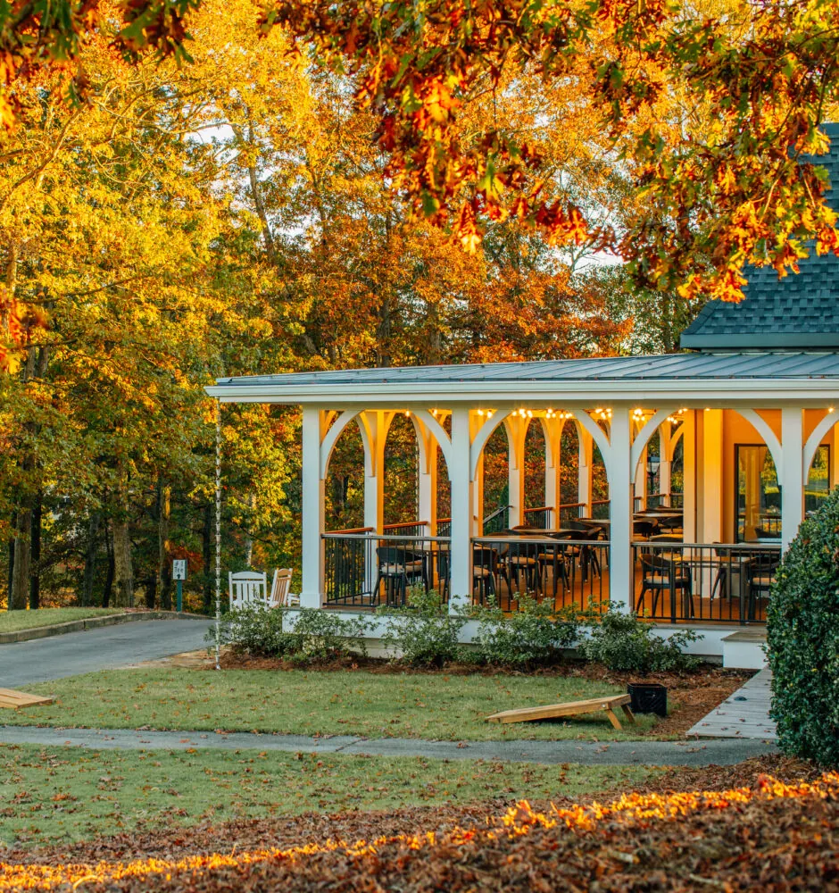 A covered outdoor porch with seating is next to a driveway and a lawn with two cornhole boards. The trees behind the porch have bright yellow and orange leaves.