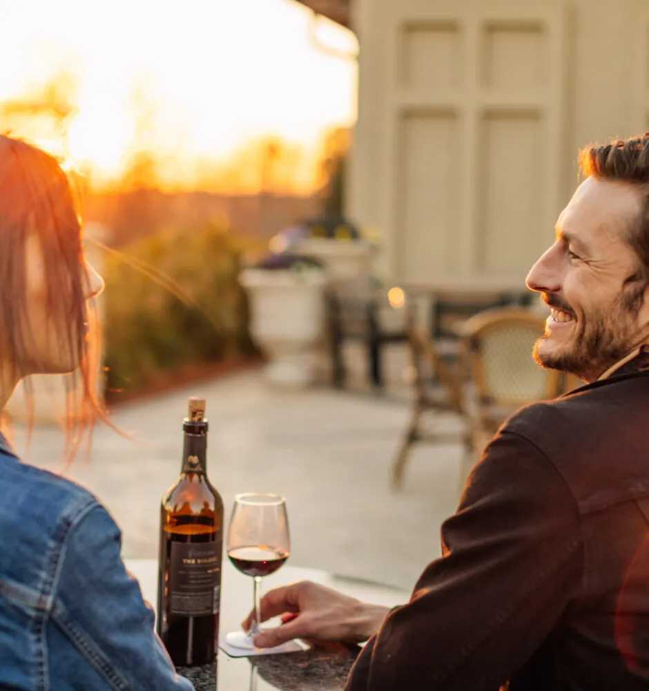 A man sits outdoors with a glass of red wine, with the sun setting behind him.