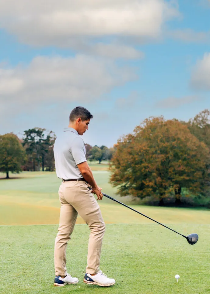 A man in a light blue shirt and tan pants is about to hit a golf ball on a green course.