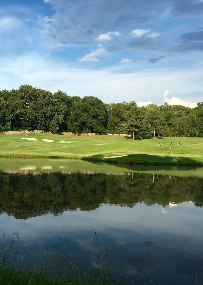 A calm lake with trees and sand traps on the opposite bank under a blue sky with scattered clouds.
