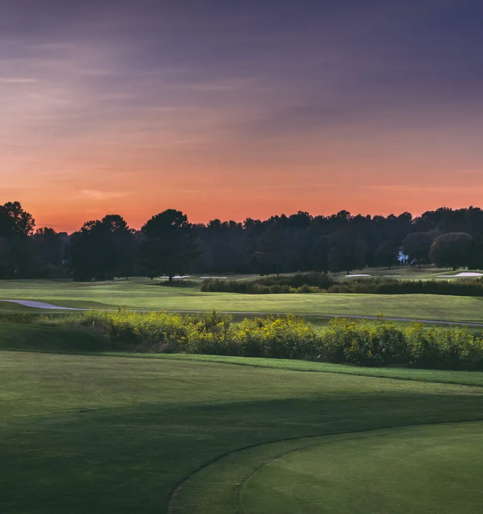 A wide view of a golf course at sunset with a mix of green grass and trees. The sun is setting, creating orange and purple clouds. Analysis