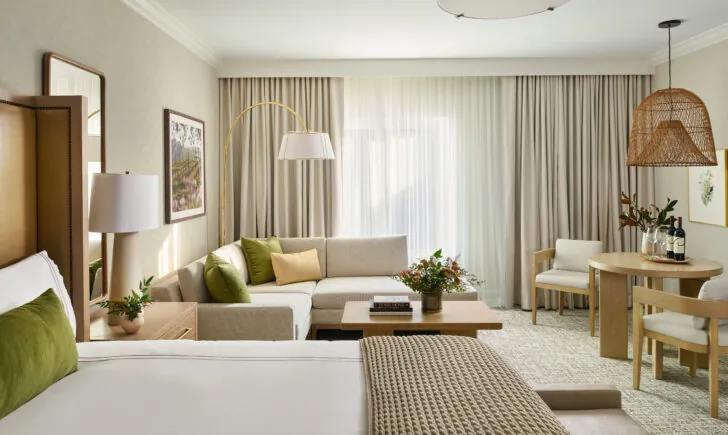 Bright suite with king bed, neutral sofa with olive green and gold pillows, wooden coffee table, rattan pendant light, and dining area with natural light