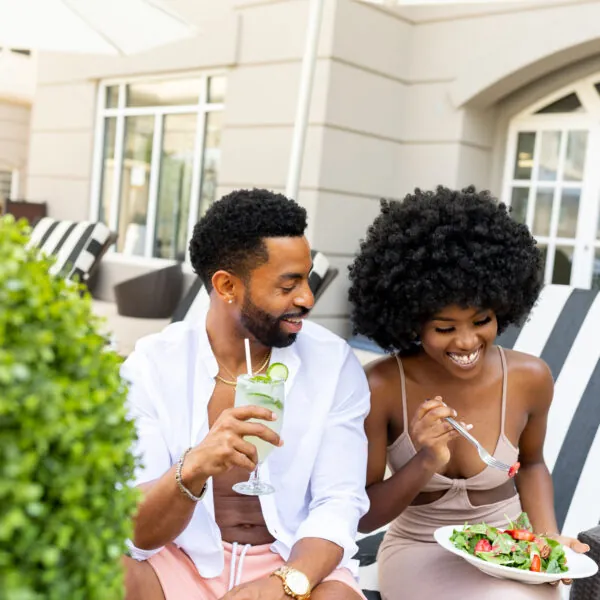 A smiling couple sits outdoors, the man holding a drink and the woman eating a salad.