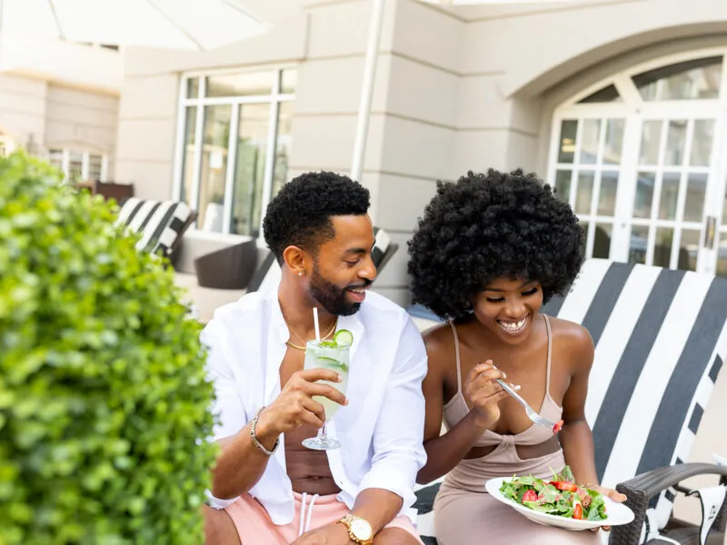 A smiling couple sits outdoors, the man holding a drink and the woman eating a salad.
