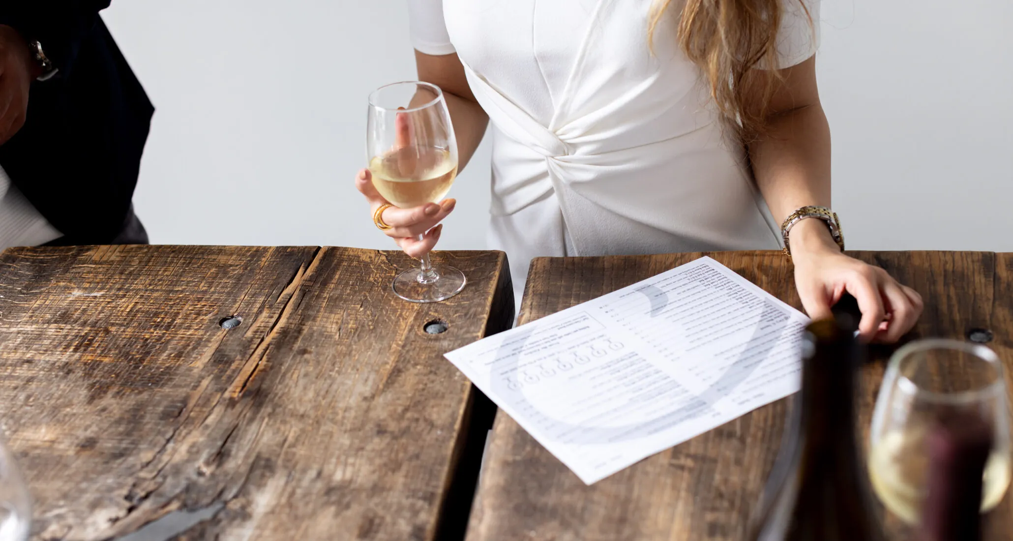 A person holds a glass of white wine while sitting at a rustic wooden table with a bottle and a piece of paper.