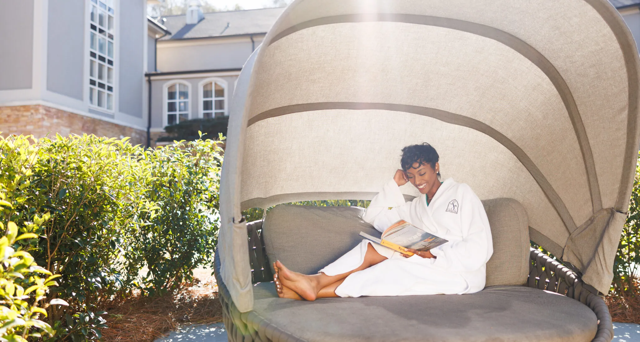 A woman in a white robe relaxes on an outdoor lounge chair, reading a book.