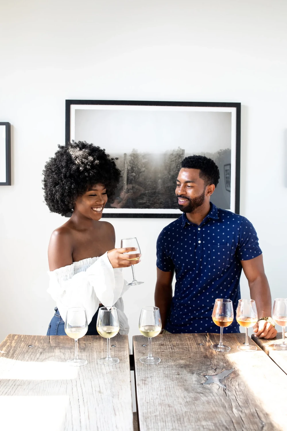 A smiling couple is standing at a wooden table. The woman, with a large afro, is wearing a white off-the-shoulder shirt and is toasting with a glass of white wine. The man, in a blue collared shirt, is looking at her and smiling. Several wine glasses are on the table in front of them.