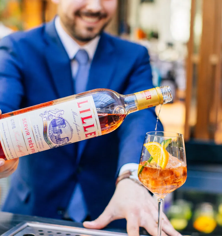 A bartender in a blue suit smiles while pouring an orange-colored liqueur from a bottle into a cocktail glass.