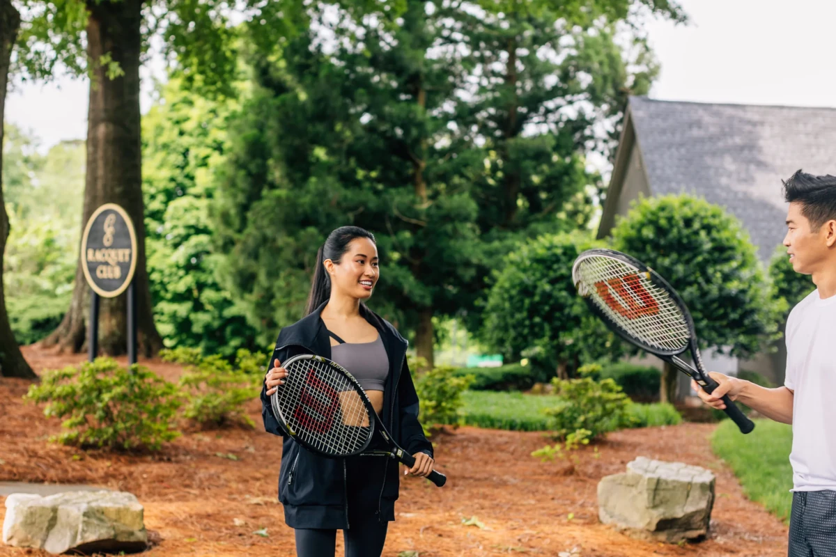 Two people hold tennis rackets and stand outdoors in a wooded area.