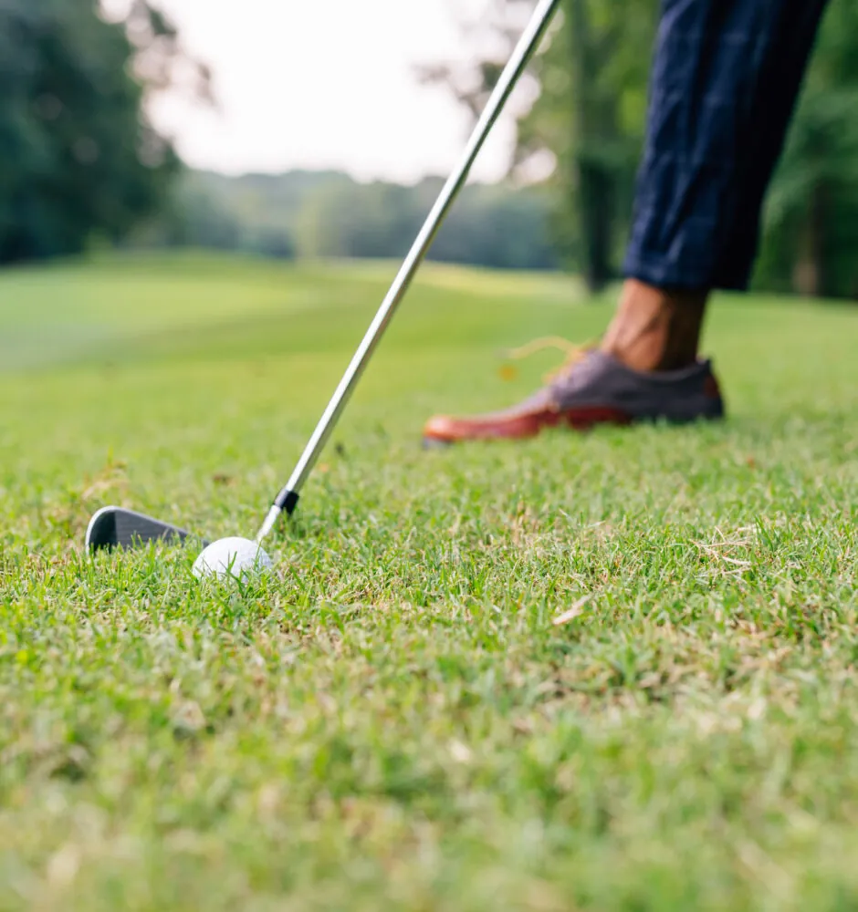 A low-angle close-up of a person's feet and a golf club poised to hit a golf ball on the grass.