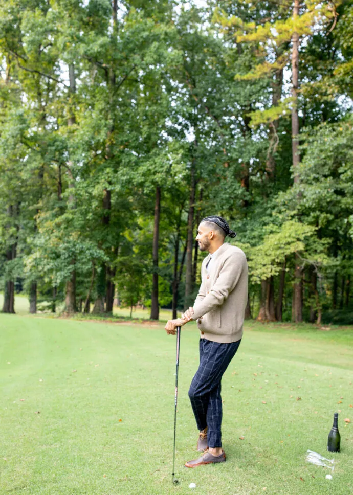 A man in a sweater stands on a golf course, holding a golf club and looking to the side.