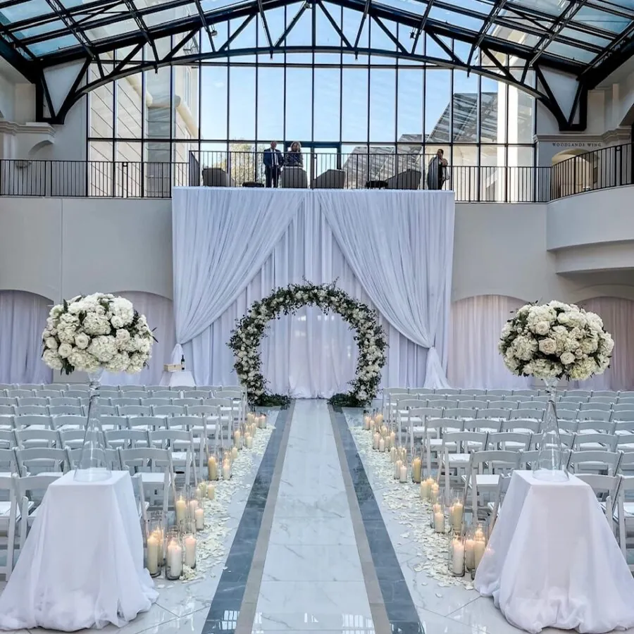 Indoor wedding ceremony with white chairs, a white aisle runner, and an arched floral arrangement.