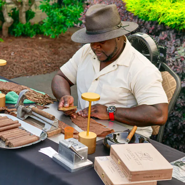 A man in a hat and apron prepares a cocktail behind a bar at an outdoor event.
