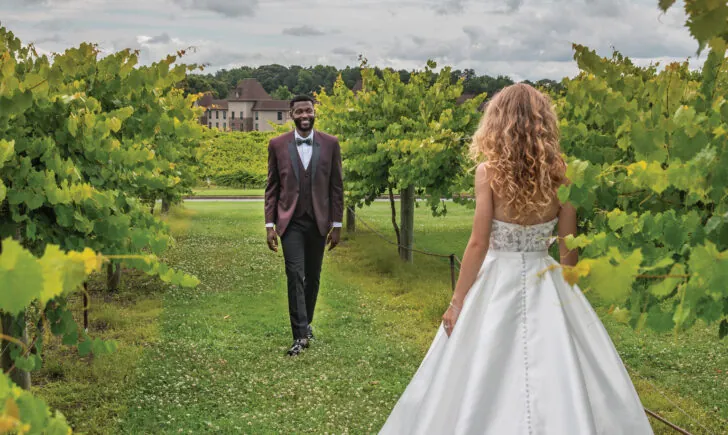 A bride in a long white dress stands in a grassy vineyard looking back at the groom, who is walking towards her.