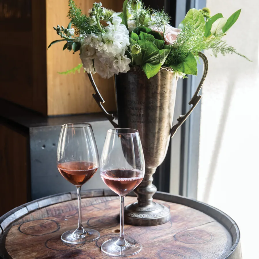 Two glasses of rosé wine on a wooden barrel, next to a flower arrangement.