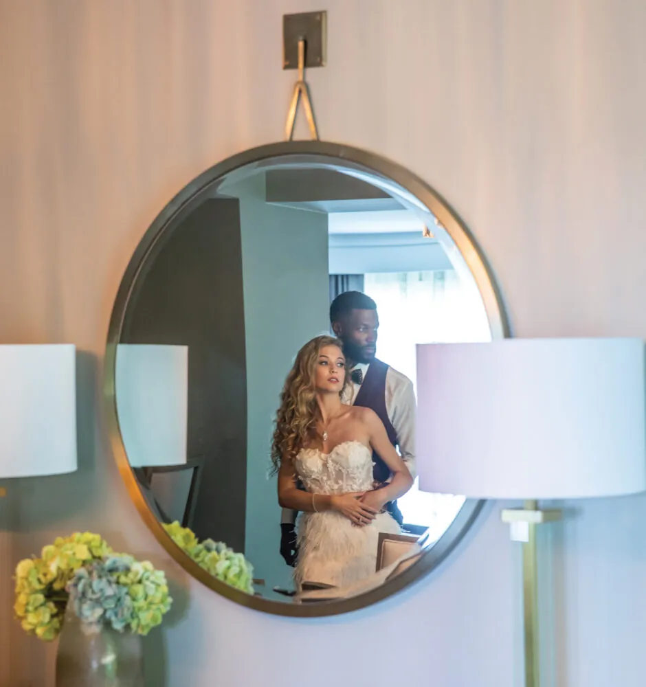 A blurry image of a couple in a mirror. The man, in a waistcoat and bow tie, is standing behind the woman, wrapping his arms around her. The woman, wearing a white strapless dress, has long, curly hair. They are both looking at the camera. The reflection shows a room with a round mirror, two lamps, and a vase of flowers.