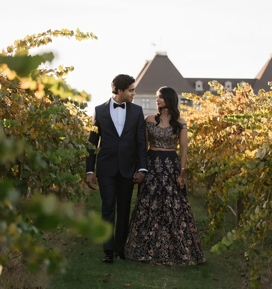 A blurry photo shows a man and a woman in formal attire walking through a vineyard. The man, on the left, is wearing a black tuxedo with a bow tie. The woman, on the right, is in a black and gold dress. They are looking at each other as they walk. In the background, a large building is visible through the grapevines.