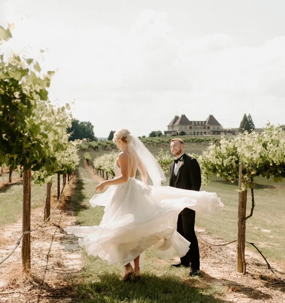A full-length photo of a bride and groom in a vineyard. The bride is spinning, her white wedding dress and veil flowing around her. The groom stands next to her, looking on. In the background, a large elegant building is visible behind the rows of grapevines.