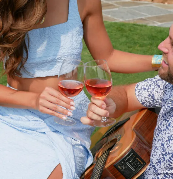 A blurred, close-up shot of a couple sitting on the grass. The woman, wearing a light blue dress, is on the left, and the man is on the right. They are clinking two glasses of rosé wine. The man is also holding a guitar.