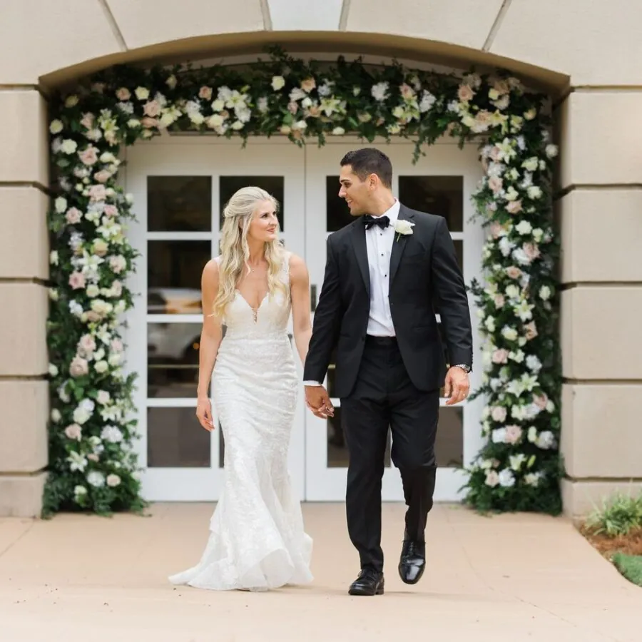 The bride and groom are walking together down a hallway with floral arches.