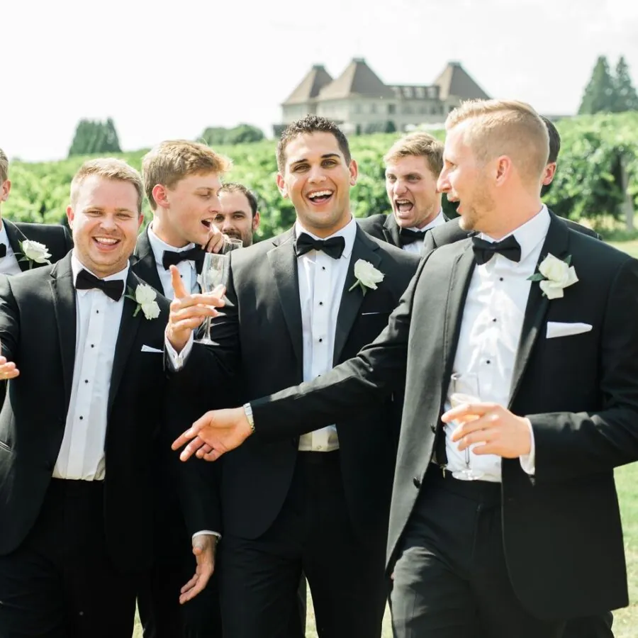 A group of men in tuxedos, including groomsmen, laughs as they walk through a grassy field.