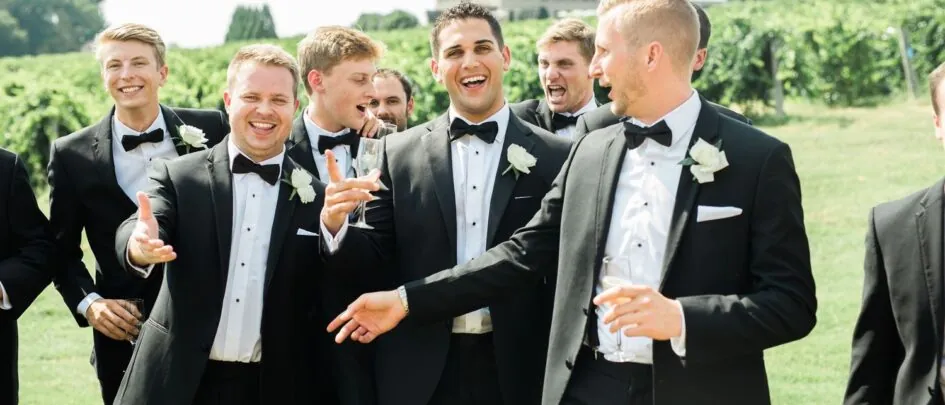 A group of men in tuxedos, including groomsmen, laughs as they walk through a grassy field.