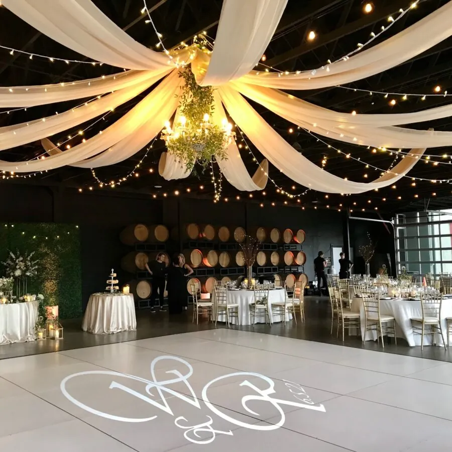 Indoor reception hall with draped fabric, string lights, and a dance floor with a monogram.