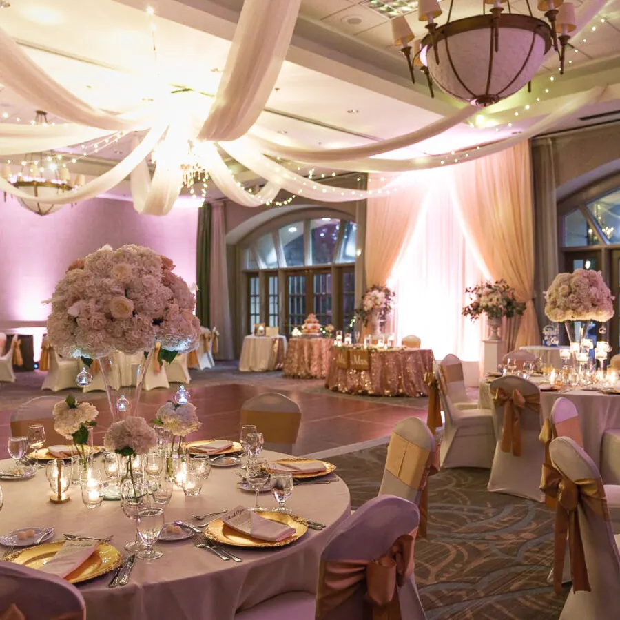 Ballroom wedding reception with draped ceilings, pink lighting, and tables decorated with gold accents and large floral centerpieces.