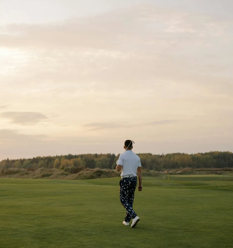 A man walks away from the camera on a green, grassy field under a cloudy, sunset sky.