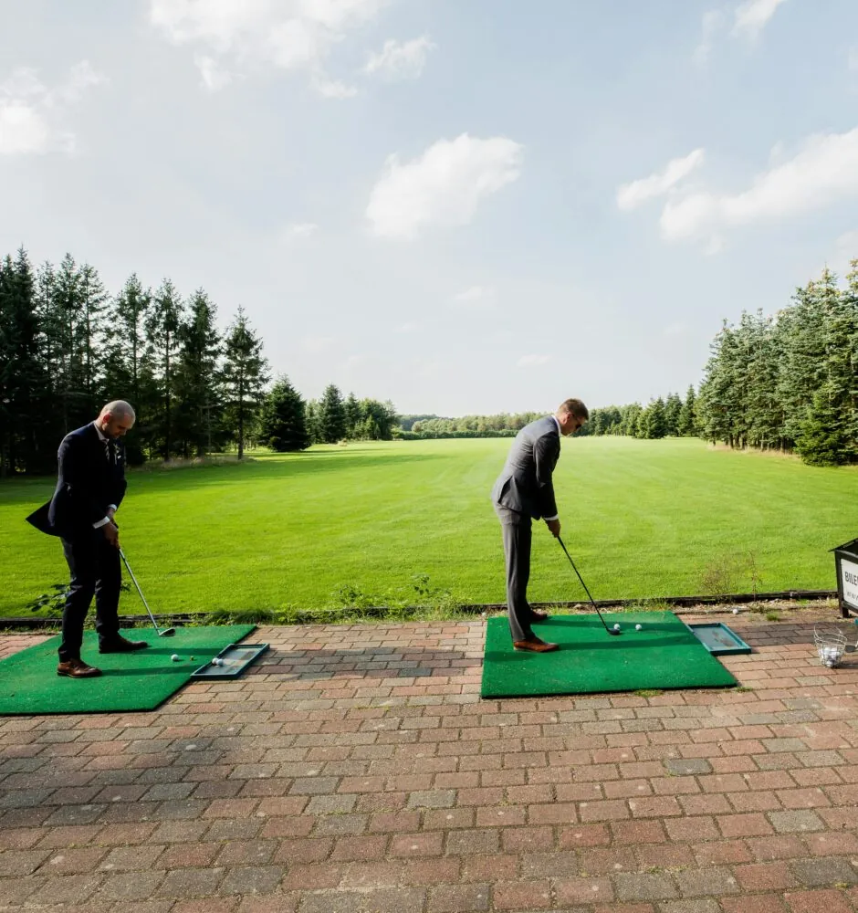 Two people in business suits practice their golf swings at a driving range.