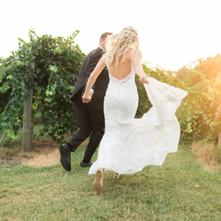 A bride and groom are walking together in a field with vineyards in the background.